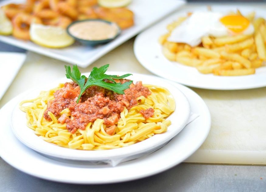 Plato de tallarines con tuco y una hoja de perejil; detrás, rabas y papas fritas con huevo frito